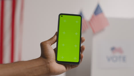 Nahaufnahme-Einer-Hand,-Die-Bei-Den-Amerikanischen-Wahlen-Ein-Mobiltelefon-Mit-Grünem-Bildschirm-Vor-Der-Wahlurne-Hält